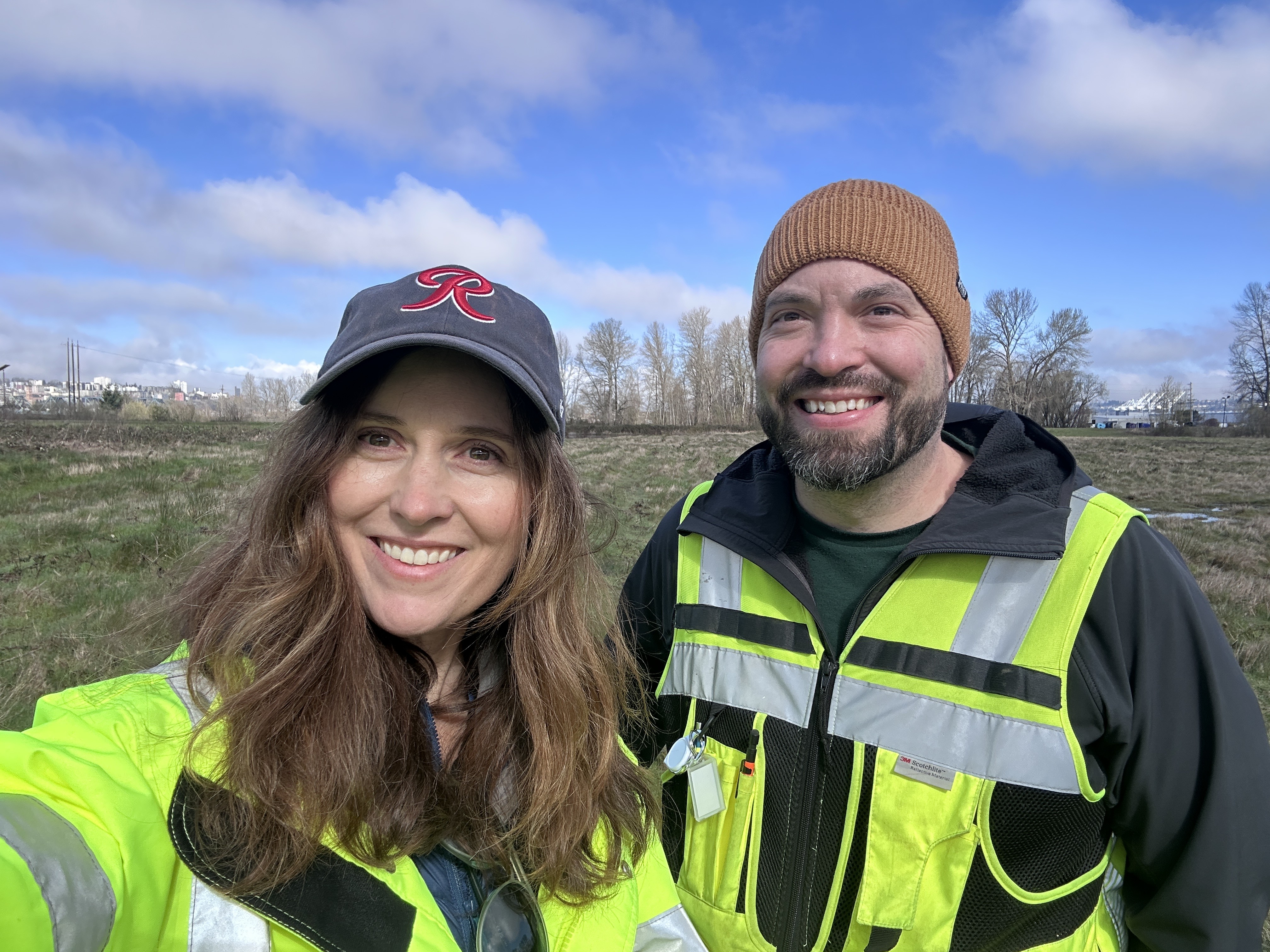 A woman (left) and man (right) in high vis vests smile in front of a blue sky.